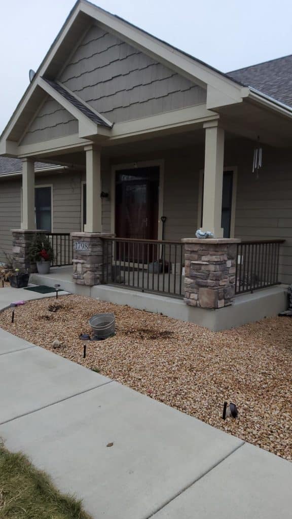 Single-story house with light brown siding, stone columns, a covered front porch, and a small garden area with gravel and flowerpots. Concrete sidewalk leads to the steps; Krumm Exteriors recently updated the gutters for added curb appeal.