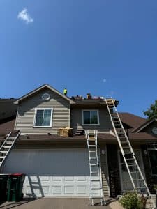 A person in a yellow shirt stands on the roof of a house under construction, with two ladders leaning against the roof and supplies for gutters and siding scattered above a white garage under a clear blue sky.