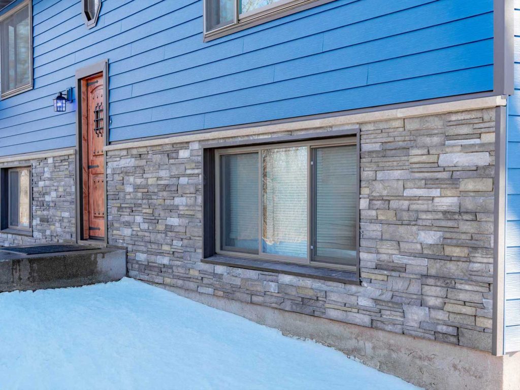 A blue house with expert siding and stonework on the lower half, featuring two large windows and a wooden front door. Snow covers the ground in front of the house.