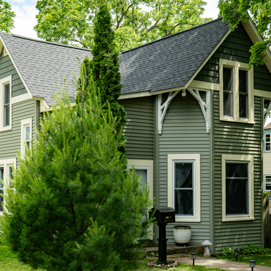 A green two-story house with white trim, surrounded by lush trees and bushes, featuring quality roofing by Krumm Exteriors, large windows, a shingled roof, and a small mailbox near the walkway.