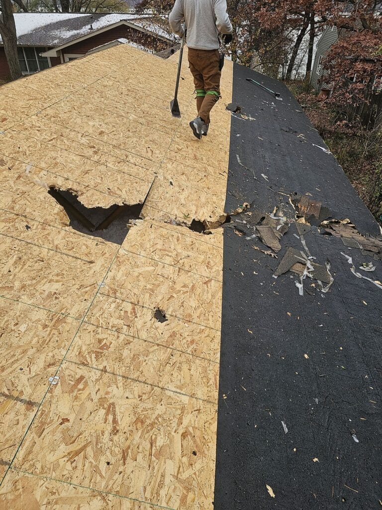 A person walks on a partially repaired roof in New Richmond WI, with one section covered in black roofing paper and the other exposed plywood. Several large holes and broken plywood sheets are visible in the unfinished area.