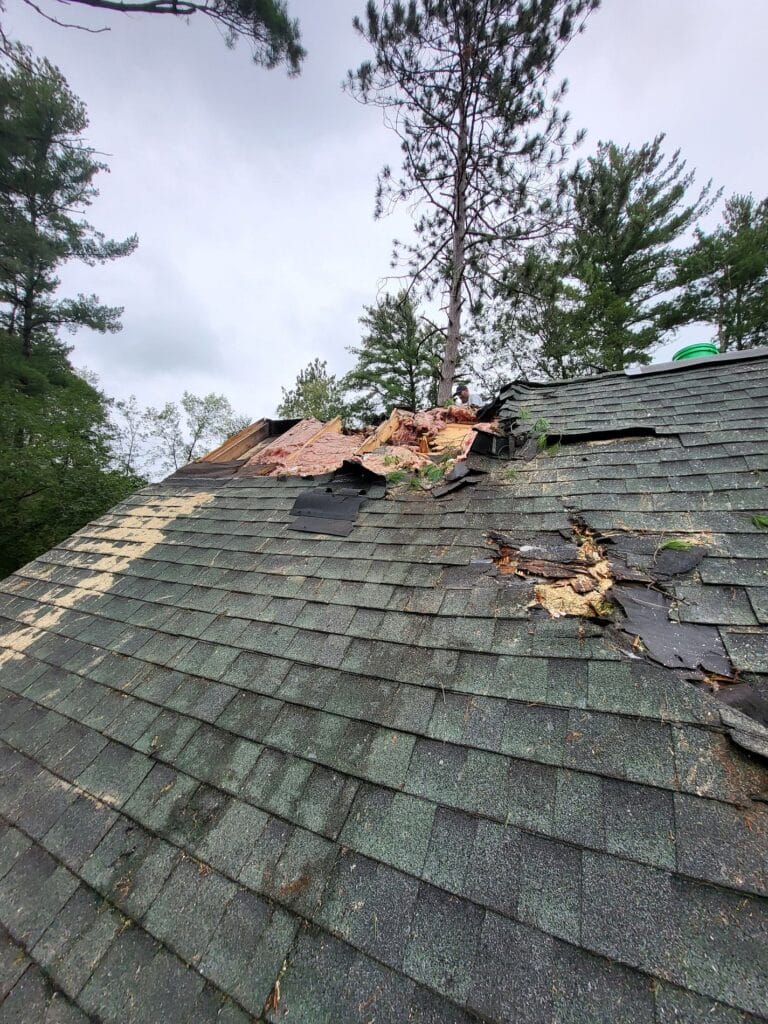 A roof with green shingles is damaged, showing exposed wood, torn shingles, and insulation. Trees surround the area under a cloudy sky—ideal for Roofing and Siding Services in Oakdale MN to restore and protect your home.