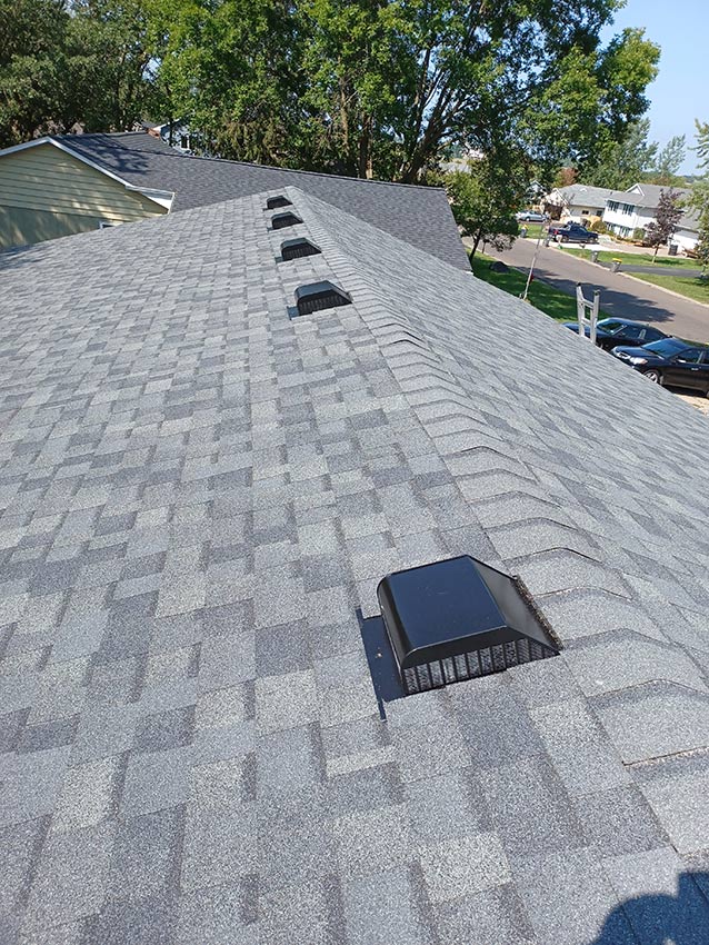 Gray asphalt shingle roof with several black vent covers evenly spaced along the ridge, surrounded by green trees and suburban houses in the background on a sunny day—showcasing quality Roofing and Siding Services in Oakdale MN.