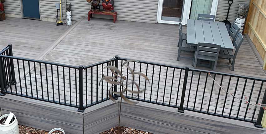 An outdoor deck with gray flooring, a dining table and chairs, a black metal railing, and a wooden privacy fence on the right—expertly finished by siding and roofing contractors in Hudson. Decorative items sit near the house’s siding in the background.