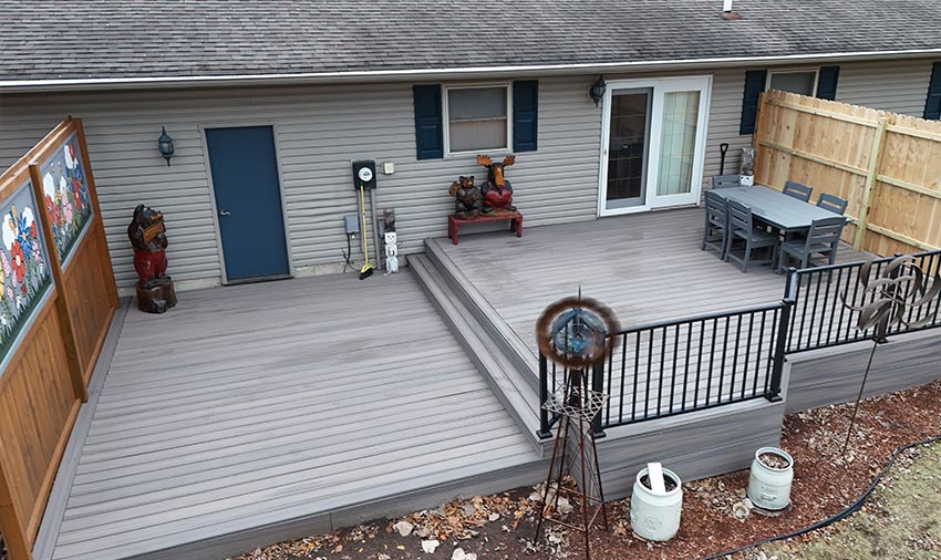 A spacious backyard deck with gray flooring, a black railing, an outdoor dining table and chairs, decorative statues, a wooden privacy screen with artwork, and a windmill ornament—perfect inspiration from top siding and roofing contractors in Menomonie.
