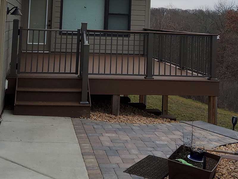 A raised wooden deck with black metal railings and stairs leads down to a stone patio. There is a planter with a small plant in the foreground, and a grassy area and trees in the background.