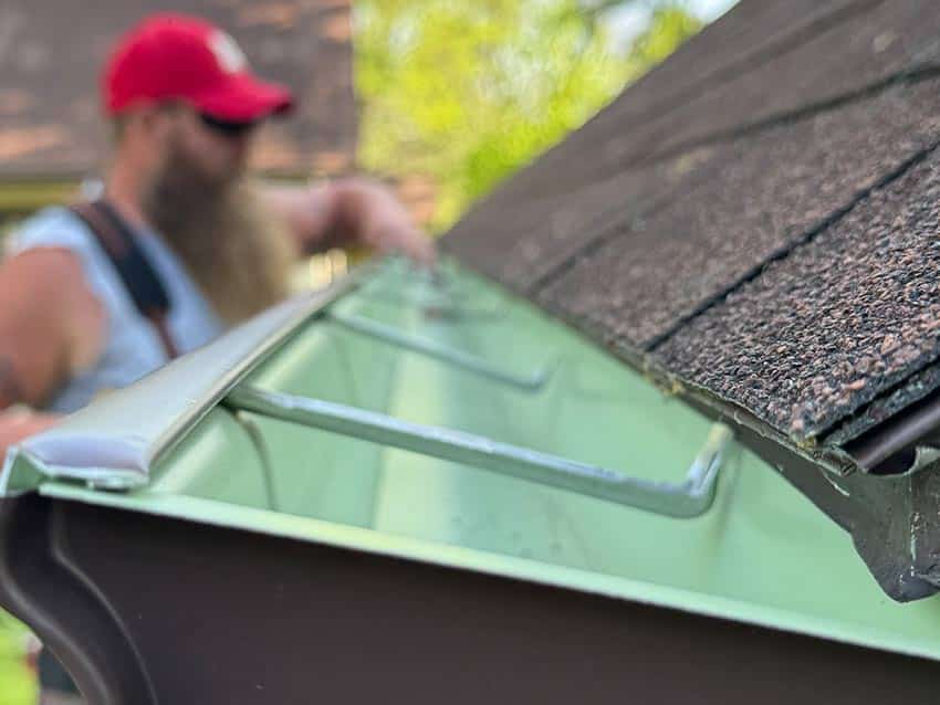 Close-up view of a house gutter along a shingled roof, with a person in the background—possibly one of the siding and roofing contractors in Eau Claire—wearing a red cap and work clothes, slightly out of focus, inspecting or cleaning the gutter.