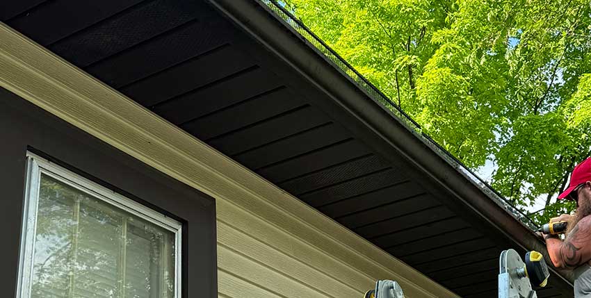 A person on a ladder installs or cleans a black gutter along the roof edge of a beige house with black trim, next to a window, under leafy green trees.