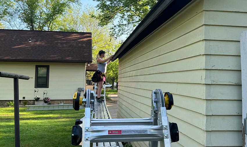 A person wearing a hat stands on a ladder, cleaning or inspecting the gutter of a beige house with yellow siding—a common task recommended by siding and roofing contractors in Menomonie. Green grass and another house are visible in the background.