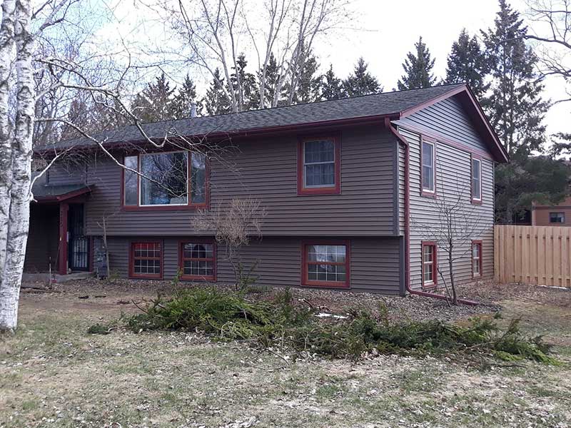 A two-story house in New Richmond with brown siding and red trim, large windows, and a black roof is surrounded by leafless trees, a wooden fence, and some fallen branches on the grass in the foreground.