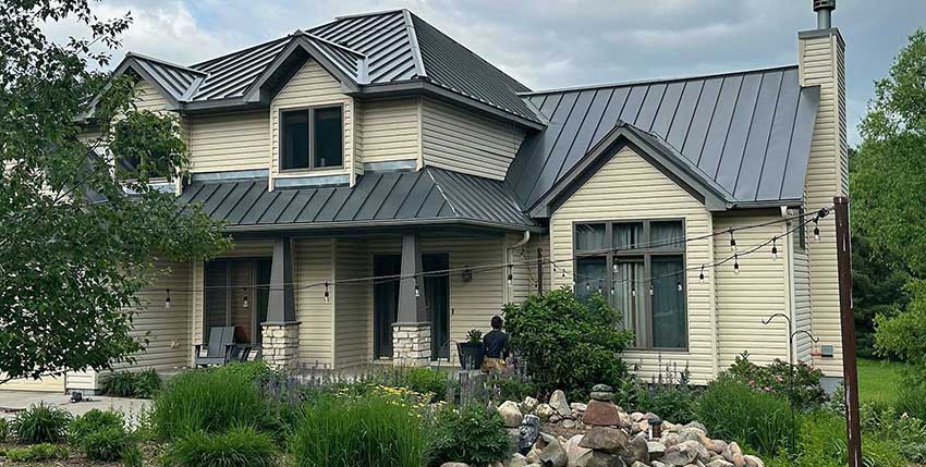 A modern beige house with a dark metal roof, large windows, and a covered porch. The front yard features lush greenery, a small rock garden, and string lights. Trees and cloudy sky are visible in the background.