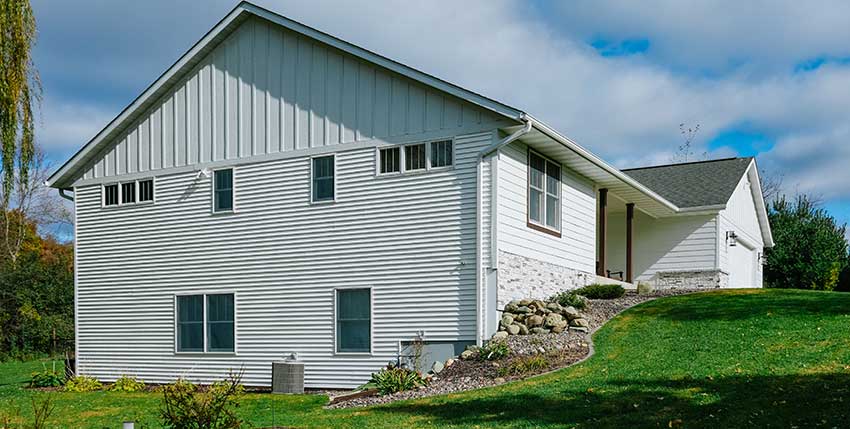 A white, single-story house with horizontal siding sits on a grassy lawn with a stone retaining wall, bushes, and trees in the background under a partly cloudy sky.