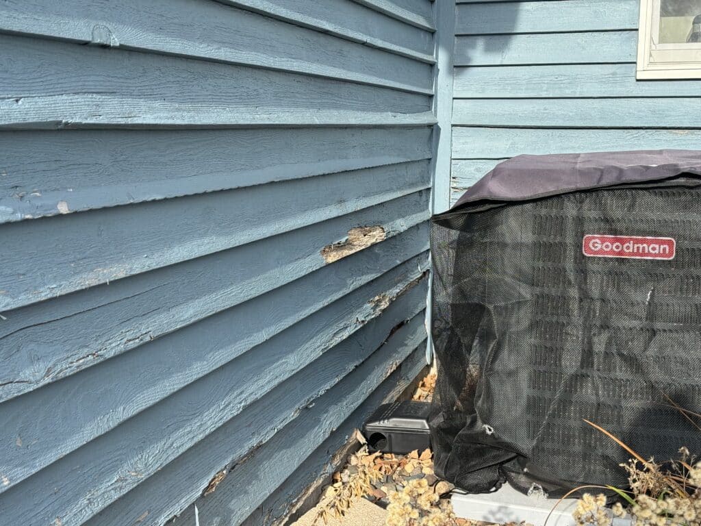 A blue wooden house exterior with visible damage and peeling on the siding near an air conditioning unit labeled “Goodman.” Dead leaves are scattered on the ground next to the unit.