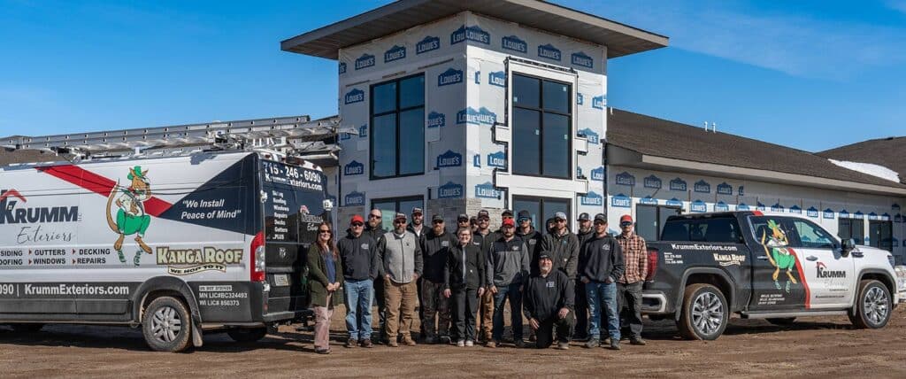A group of people wearing work clothes stand in front of a partially constructed building, flanked by two branded Krumm Exteriors vehicles under a clear blue sky.