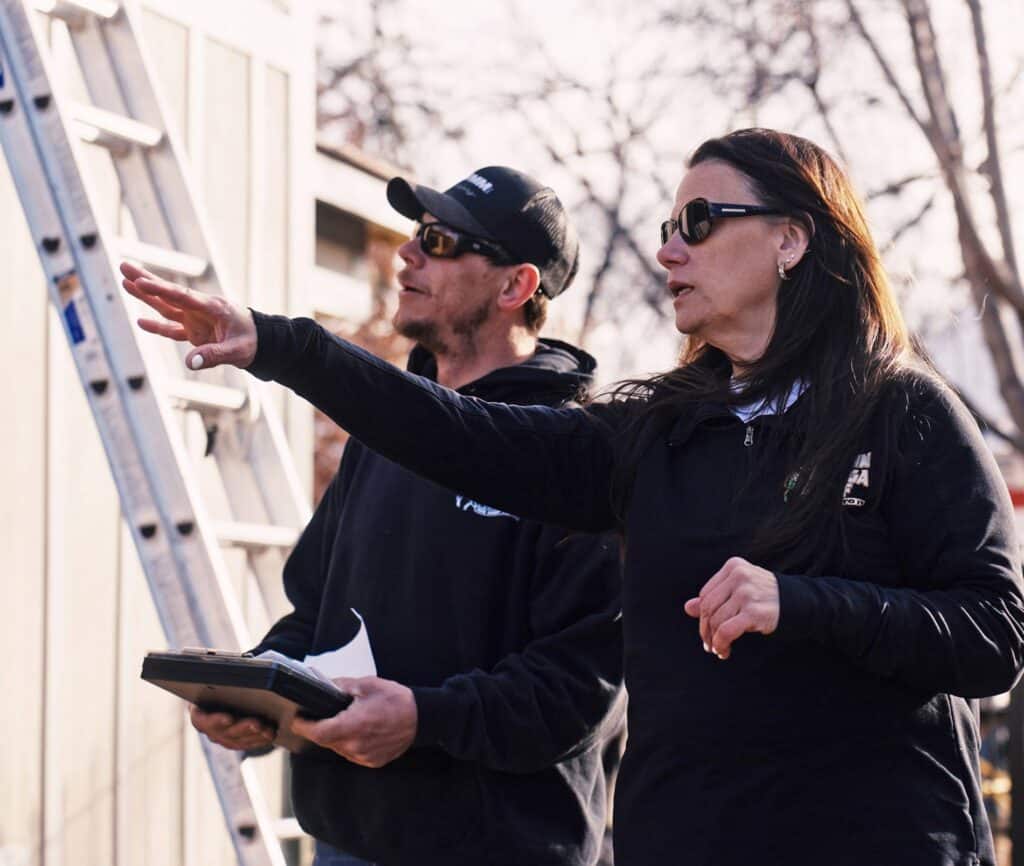 Two people wearing dark clothing and sunglasses stand outdoors near a ladder. One holds a clipboard, while the other points ahead, appearing to discuss or inspect something at a work site. Leafless trees are visible in the background.