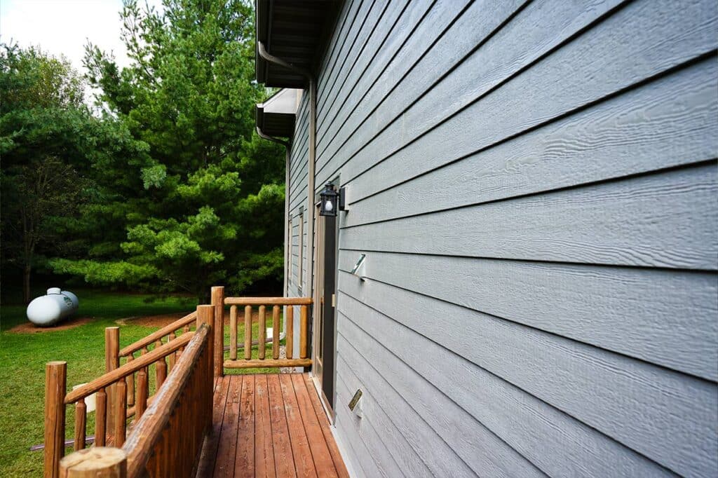 A wooden deck with a log railing runs alongside a gray house exterior with LP Smart Siding. Lush green trees are visible in the grassy yard in the background.