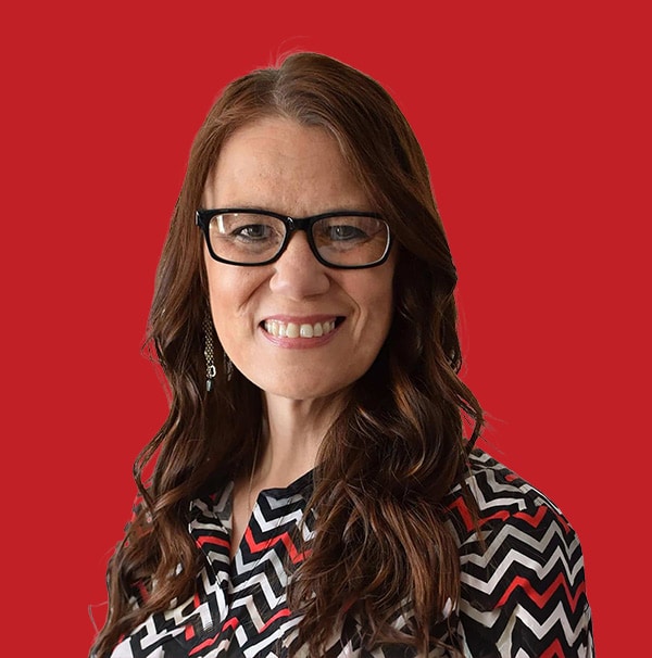 A woman with long brown wavy hair and glasses smiles in front of a solid red background. She is wearing a black, white, and red chevron-patterned top.