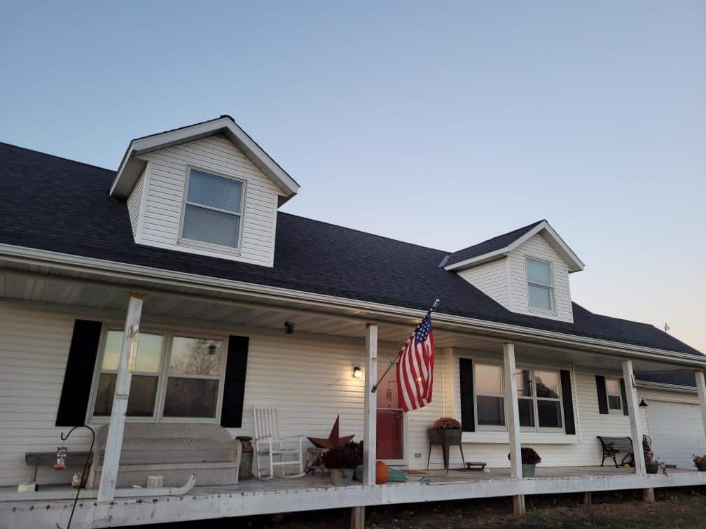 A white house with a front porch, two dormer windows, an American flag, porch swing, benches, and autumn decorations at dusk under a clear sky—showcasing quality roofing and siding and Gutter services in Oakdale MN.