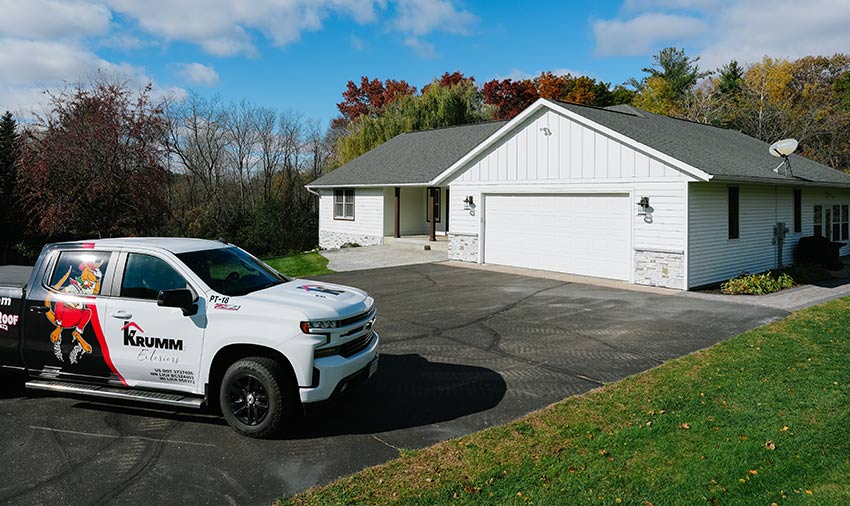 A white pickup truck with “Krumm Exteriors” branding, siding and roofing contractors in Menomonie, is parked in the driveway of a modern, single-story house with a large garage, surrounded by trees under a partly cloudy sky.