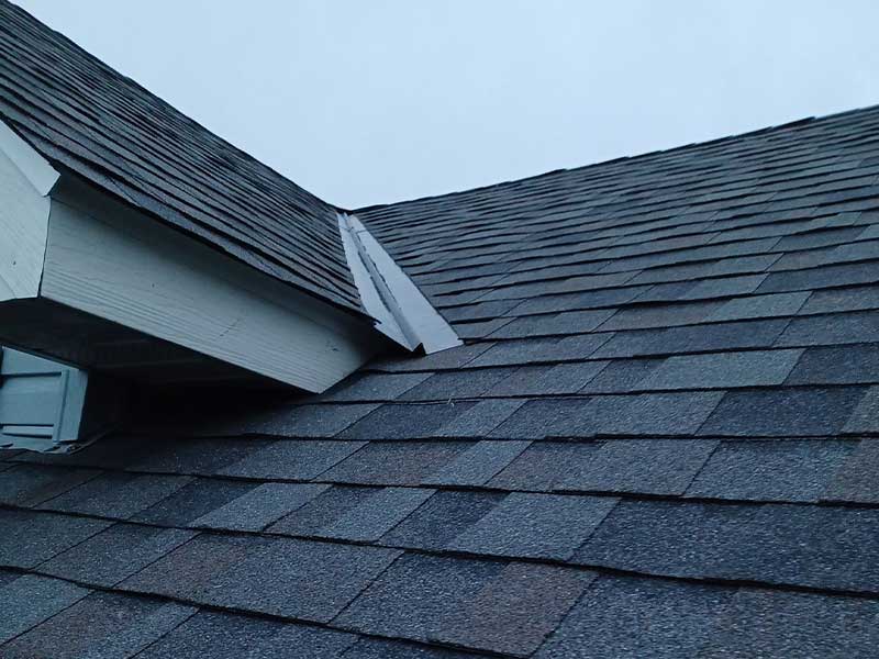 Close-up view of a New Richmond house roof with gray asphalt shingles, featuring a small dormer and a sloped valley section meeting at an angle, under a cloudy sky.