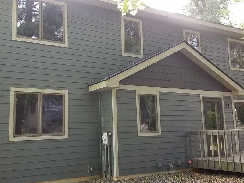 A two-story house with gray horizontal siding, beige trim, multiple windows, a small porch with a railing, and gravel along the foundation. Trees are visible in the background.