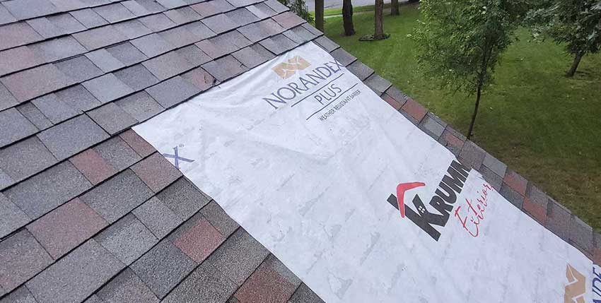 A house roof under construction with asphalt shingles partially installed and a white Norandex Plus and Krumwiede Exteriors branded underlayment visible on the exposed section. Trees and grass are in the background.