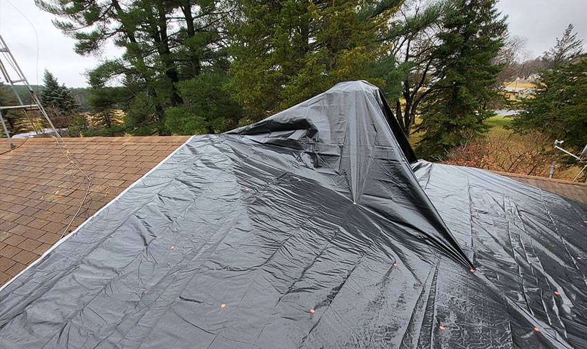 A black plastic tarp covers a roof, held down with orange fasteners—an example of the temporary solutions siding and roofing contractors in Menomonie might use, surrounded by trees and an overcast sky in the background.