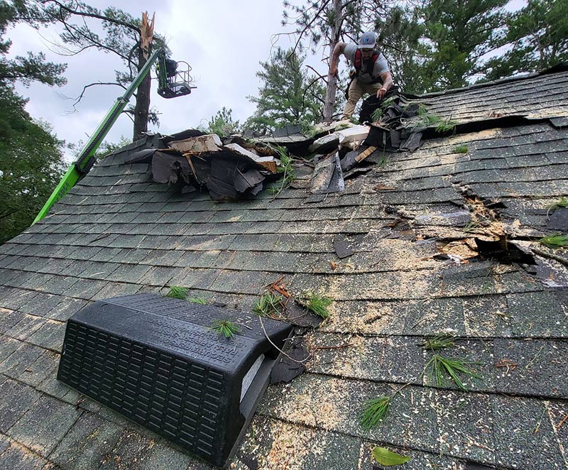 A worker repairs a roof with heavy damage and broken shingles, likely from a fallen tree; debris and pine needles are scattered, and a large vent is attached to the roof. A lift is visible in the background among the trees.