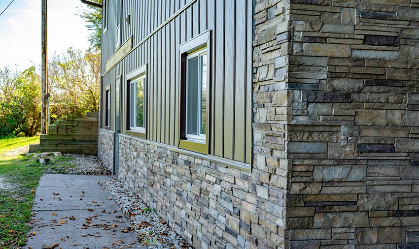 A modern house exterior with gray stone veneer siding and large windows, crafted by expert siding and roofing contractors in Menomonie, sits next to a concrete walkway bordered by rocks and lush greenery in the background.