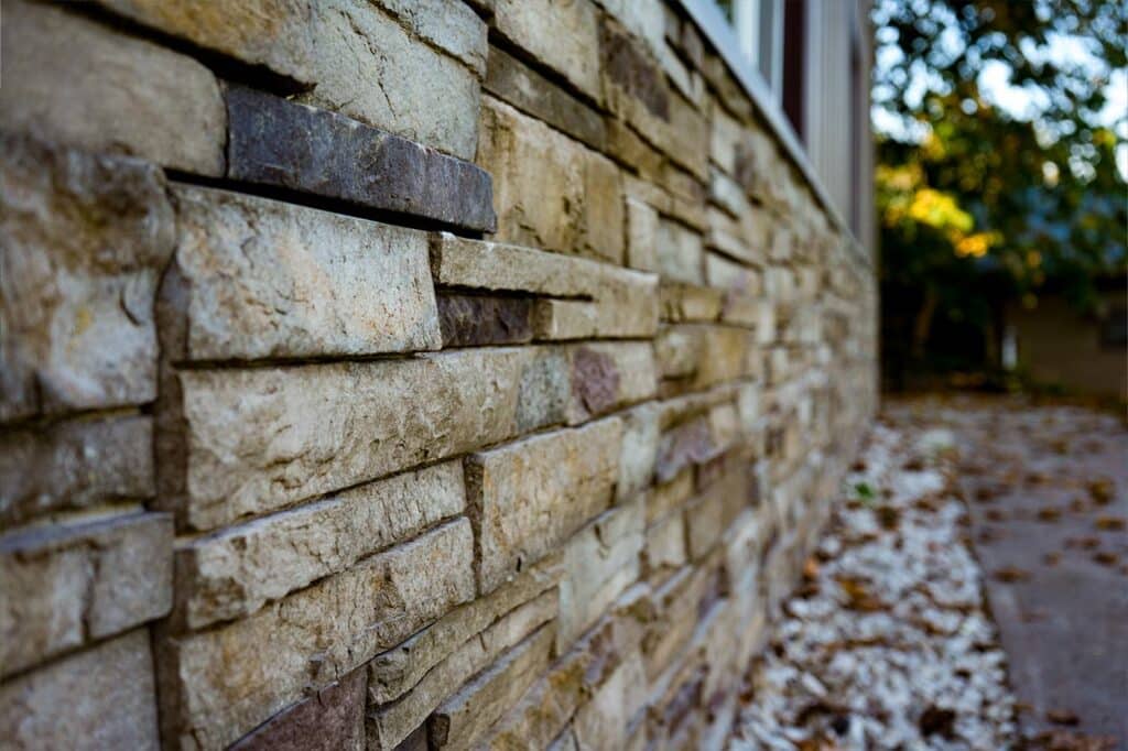 Close-up of a stone wall with professionally installed Versetta Stone. The textured wall runs alongside a walkway bordered by white rocks and fallen leaves.