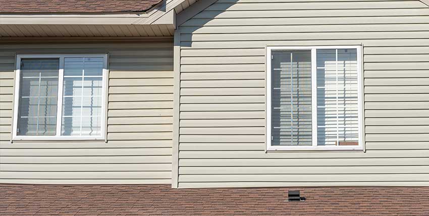 Beige vinyl siding on a house exterior with two white-framed windows, both featuring white blinds. The brown shingled roof is visible at the bottom of the image.