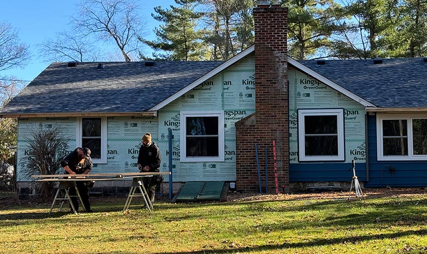 Two people work on wood at saw horses in front of a house under renovation, with siding and roofing contractors in Menomonie updating the exterior—part is covered in building wrap, part in blue siding. A brick chimney stands in the center, with trees behind.