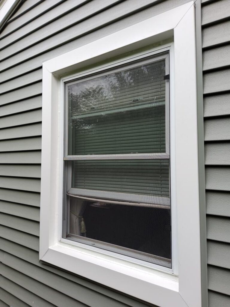 A double-hung window with white trim on a gray, horizontally-sided house in Oakdale MN. The window is partially open with the lower sash raised, showing a green window blind and some trees reflected in the glass—Roofing and Siding Services in Oakdale MN.