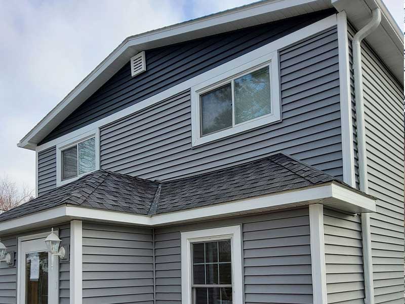 Two-story house with gray vinyl siding, white trim, and white-framed windows. The roof has gray shingles, and a small porch roof extends over the front door. A white lamp is mounted near the entrance.