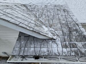 A snow-dusted house roof with black heating cables arranged in loops to prevent ice dams. The white gutters below are mostly clear, and the shingles are partially covered with a thin layer of snow.