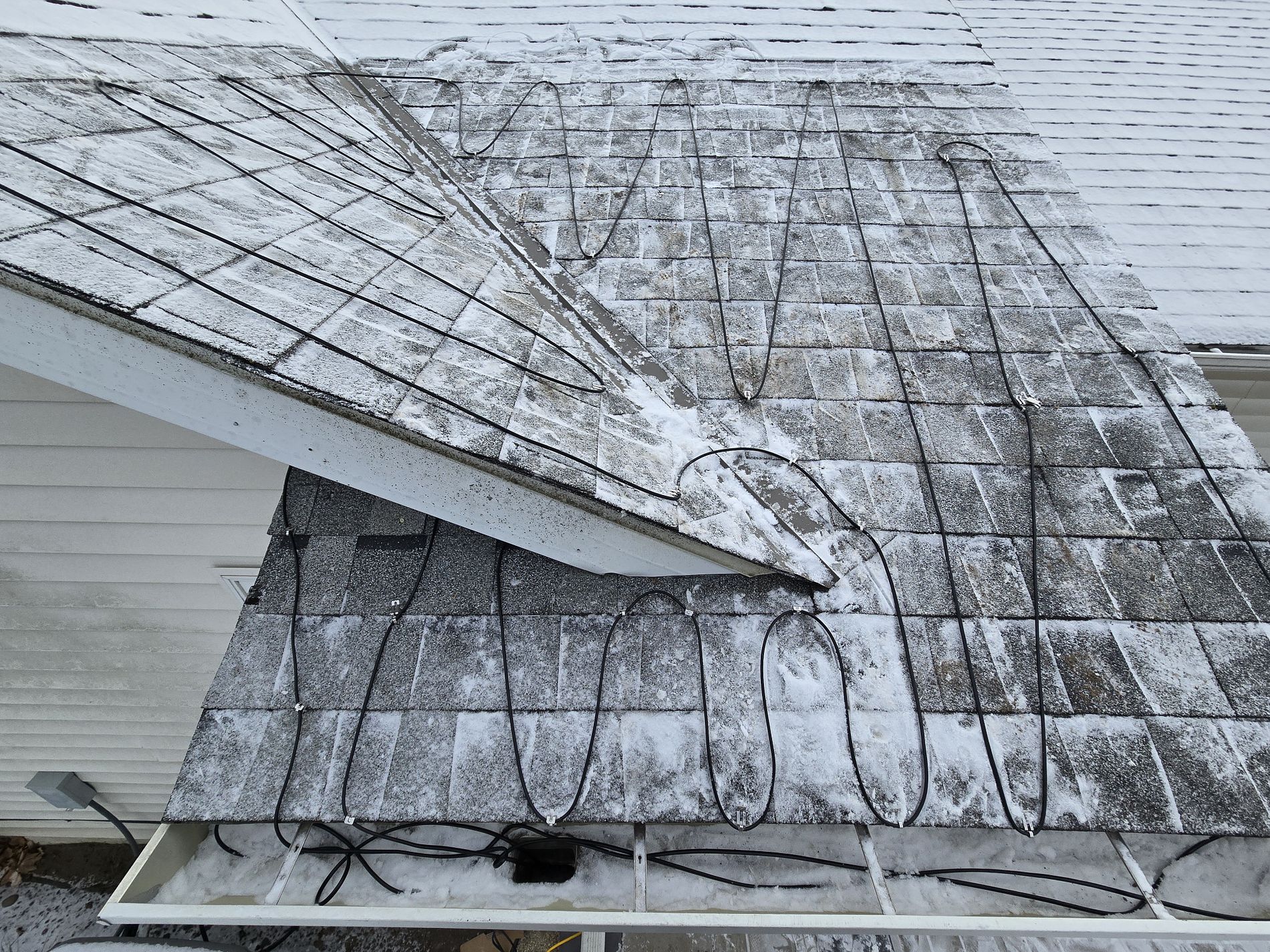 A snow-dusted house roof with black heating cables arranged in loops to prevent ice dams. The white gutters below are mostly clear, and the shingles are partially covered with a thin layer of snow.