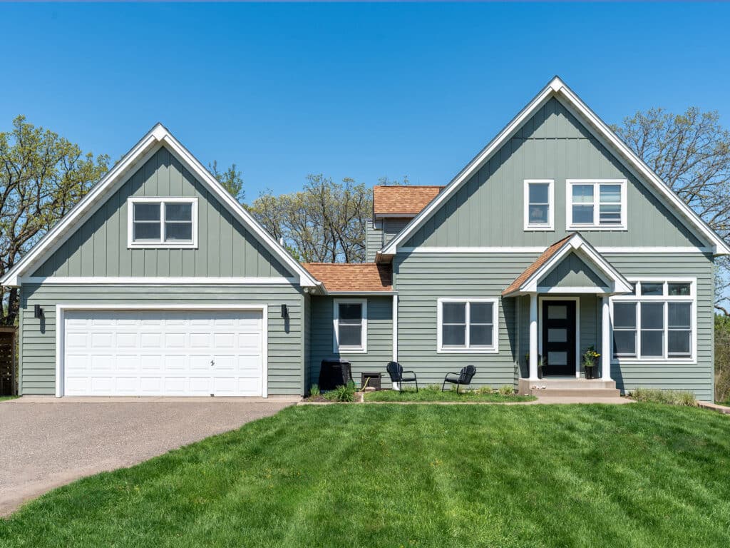 A modern two-story house with professionally installed new light green siding, white trim, a double garage, black front door, chairs on the porch, and a well-kept green lawn under a clear blue sky.