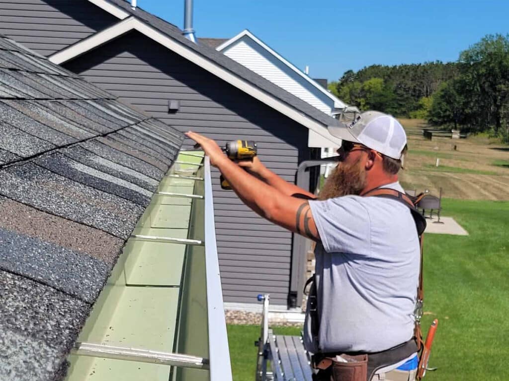 A man wearing a cap and harness, uses a power drill to install or repair a rain gutter on the edge of a house roof on a sunny day.
