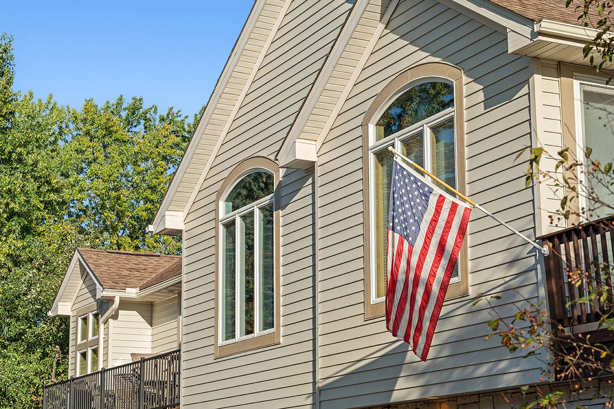 A house with light beige seamless steel siding and large arched windows displays an American flag on a pole. Trees surround the home, and the sky is clear and blue.