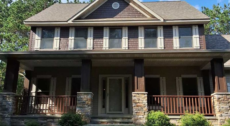 Two-story house in Hudson WI with brown siding and white trim, large covered front porch with wooden railings, stone pillars, and neatly trimmed green shrubs and lawn in front.